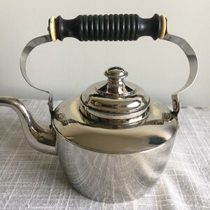 Puede incluir: Una tetera de plata con un mango de madera negro y un acabado brillante. La tetera tiene un cuerpo redondeado y un pico en el lateral.