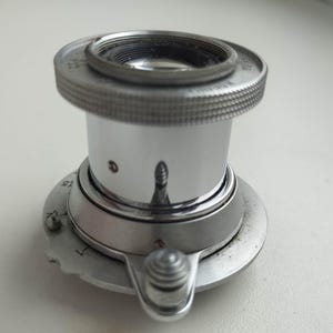 Puede incluir: Una lupa plateada de estilo vintage, con un anillo moleteado alrededor de la lente. Tiene una base circular con un peque&ntilde;o bot&oacute;n de ajuste y una escala. La superficie reflectante muestra un reflejo distorsionado.