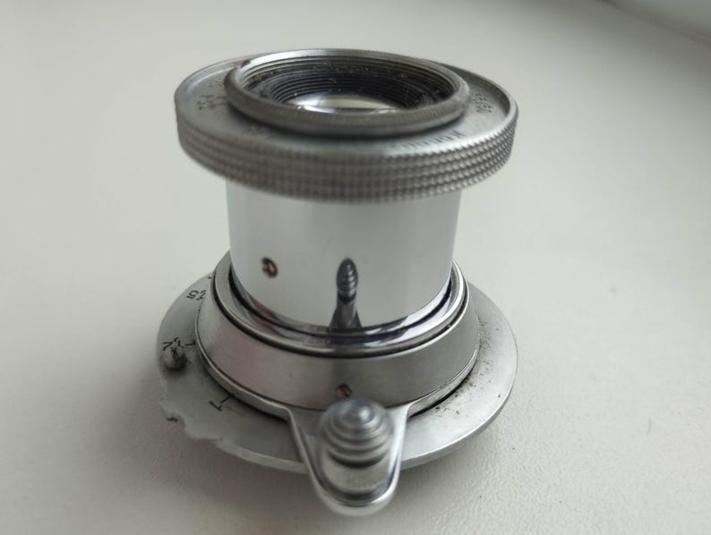 Puede incluir: Una lupa plateada de estilo vintage, con un anillo moleteado alrededor de la lente. Tiene una base circular con un peque&ntilde;o bot&oacute;n de ajuste y una escala. La superficie reflectante muestra un reflejo distorsionado.