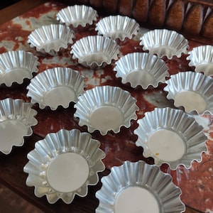 Puede incluir: Una colección de 18 moldes para hornear redondos, acanalados y de color plateado. Los moldes están dispuestos en un patrón aleatorio sobre una superficie de mármol rojo y blanco.