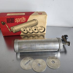 Könnte beinhalten: Eine Vintage-Keksmaschine aus Metall mit einer rot-beigen Schachtel mit der Aufschrift "KAK sprits". Die Maschine hat einen zylindrischen Körper, einen Griff und mehrere austauschbare Scheibenformen. Die Schachtel zeigt eine Abbildung der Maschine und der Keksformen.