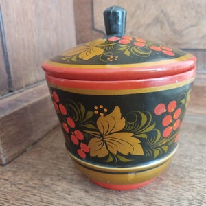 Op de afbeelding: Een kleine, ronde houten container met een deksel. De container is zwart geverfd met gouden en rode bloemmotieven. Het deksel heeft een zwarte knop.