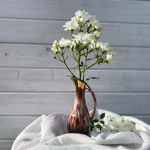 May include: A bouquet of white roses in a brown and white speckled ceramic vase with a gold rim. The vase is sitting on a white fabric surface.