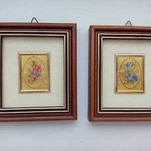 May include: Two framed pictures with gold leaf backgrounds and floral designs. The picture on the left features a red flower with green leaves. The picture on the right features a blue flower with green leaves.