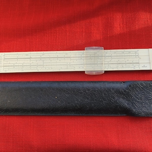 May include: A vintage slide rule with a clear cursor and a black leather case. The slide rule is off-white with black markings and numbers. The case is rectangular and has a textured surface. The background is a solid red.