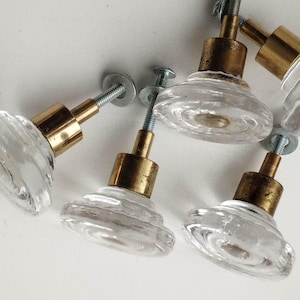 May include: A collection of six clear glass cabinet knobs with a ridged design. Each knob features a brass-colored base and a silver-colored screw for mounting. The knobs are arranged on a white surface.