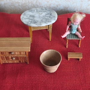 May include: Miniature dollhouse furniture set including a round table with a marble top, a small wooden dresser, a chair with a doll, a small table, and a ceramic pot. The items are arranged on a red surface.