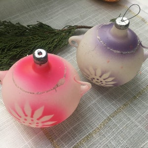 May include: Two vintage glass Christmas baubles with a white snowflake design. One bauble is pink and the other is purple.