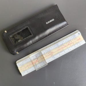 May include: A white and beige slide rule with a clear plastic cursor, measuring approximately 25 cm in length, accompanied by a black rectangular Casio branded carrying case with a small transparent window and a circular speaker grille.