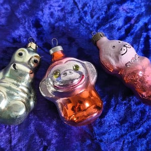 Puede incluir: Tres adornos navideños vintage con forma de animales. Los adornos incluyen un hipopótamo plateado, un mono con cuerpo naranja y una criatura rosa y blanca. Cada adorno tiene un lazo de metal para colgar.