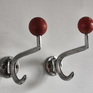 Puede incluir: Dos percheros de metal vintage. Cada gancho tiene un acabado plateado, un diseño curvo y una bola de madera roja en la parte superior. Los ganchos están montados en una pared blanca, ofreciendo una solución de almacenamiento clásica.