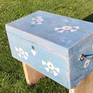 Puede incluir: Una caja de madera azul con una tapa con bisagras y asas de metal. La caja está decorada con flores blancas y hojas verdes. La caja está sentada sobre un soporte de madera.