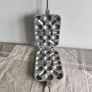 Könnte beinhalten: Offenes, vintage Waffeleisen aus Gusseisen in Pilzform. Das Scharnier-Eisen ist silberfarben und hat einen langen Griff. Das Waffeleisen ist geöffnet und zeigt die Formen für die Waffeln. Das Eisen liegt auf einer karierten Oberfläche.