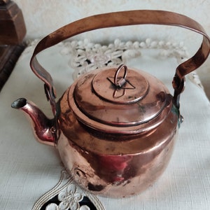 Puede incluir: Una tetera de cobre vintage con tapa abatible y asa curva. La tetera tiene un acabado deslustrado y muestra signos de antigüedad.