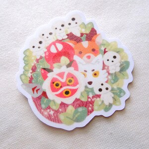 Pode incluir: Um autocolante recortado com uma ilustra&ccedil;&atilde;o colorida de v&aacute;rias criaturas e plantas da floresta. O design inclui uma m&aacute;scara vermelha e branca, raposas, esp&iacute;ritos brancos e um cogumelo, tudo rodeado por folhas verdes e bagas vermelhas.