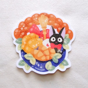 Pode incluir: Um autocolante com um gato preto de desenho animado com uma gravata borboleta vermelha, rodeado por uma tigela azul cheia de doces laranja, uma vassoura e um la&ccedil;o vermelho. O autocolante tem uma borda ondulada laranja com brilhos e folhas verdes.