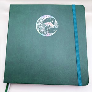Peut inclure: Un journal à couverture rigide vert foncé avec un motif de croissant de lune holographique représentant un champignon et un papillon. Le journal a un marque-page en ruban turquoise.