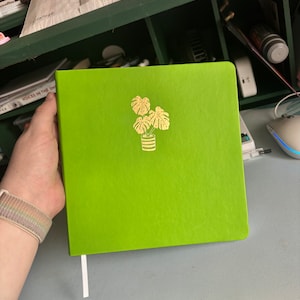 Könnte beinhalten: Ein leuchtend grünes Hardcover-Journal mit einer goldenen Folienillustration einer Topfpflanze mit drei Blättern. Das Journal hat ein weißes Band-Lesezeichen.