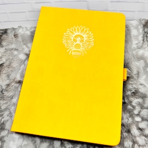 Op de afbeelding: Een geel notitieboek met een wit ontwerp van een zonnebloem en een bij op de voorkant.