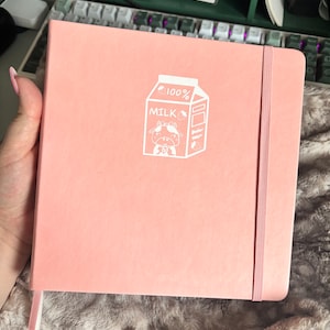Può includere: Un quaderno rosa con un design a forma di cartone del latte bianco che presenta una mucca dei cartoni animati e il testo "100% LATTE". Il quaderno ha una chiusura con elastico bianco e un segnalibro a nastro bianco.