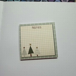 May include: A square sticky note with a grid pattern and the word "Notes" at the top. The note features three Christmas tree illustrations. The background is a dotted paper.