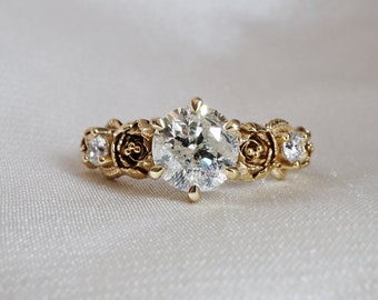 May include: A gold ring with a large, round, clear diamond in the center. The band is decorated with three small, clear diamonds and three rose-shaped designs.