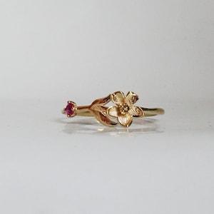 以下が含まれることがあります： 繊細な花のデザインと小さなルビーの宝石が付いた金の指輪。花は細部まで作り込まれており、質感のある仕上げになっています。