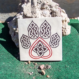 Puede incluir: Un azulejo de cerámica blanco con un diseño de nudo celta negro y rojo en forma de huella de pata. El diseño presenta un corazón en el centro de la huella de pata.