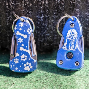 Puede incluir: Dos llaveros de metal azul con un diseño de perro. Un llavero tiene un patrón de huellas de patas y huesos, el otro tiene una silueta de perro. Ambos llaveros tienen un anillo plateado.
