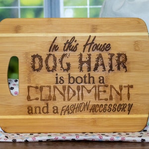 Könnte beinhalten: Ein braunes, rechteckiges Schneidebrett aus Holz mit einem Griff. Auf dem Brett ist der Text "In this house dog hair is both a condiment and a fashion accessory" eingraviert.