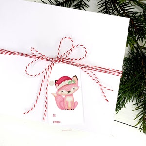 May include: A white gift package tied with red and white twine, featuring a tag with a pink fox wearing a Santa hat. The tag includes "to:" and "from:" fields, perfect for holiday gifting. Green pine branches are in the background.