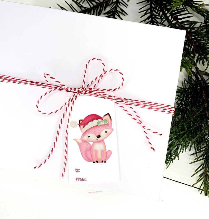 May include: A white gift package tied with red and white twine, featuring a tag with a pink fox wearing a Santa hat. The tag includes "to:" and "from:" fields, perfect for holiday gifting. Green pine branches are in the background.