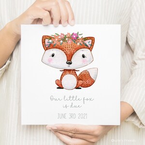 May include: A white card with a cute cartoon fox wearing a flower crown. The text on the card reads "Our little fox is due June 3rd 2021".