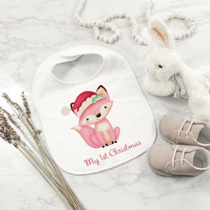 May include: A white baby bib with a pink fox wearing a Santa hat and the text "My 1st Christmas." A white stuffed bunny, a pair of beige baby shoes, and dried lavender are also present.