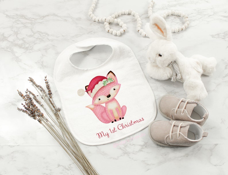 May include: A white baby bib with a pink fox wearing a Santa hat and the text "My 1st Christmas." A white stuffed bunny, a pair of beige baby shoes, and dried lavender are also present.