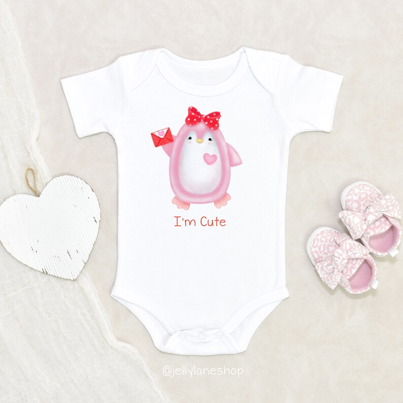 Op de afbeelding: Witte baby romper met een roze pingu&iuml;n die een rode envelop vasthoudt en een rode strik draagt. De pingu&iuml;n heeft een klein roze hart op zijn borst. De tekst "I'm Cute" is onder de pingu&iuml;n gedrukt.