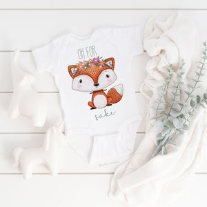 May include: White baby onesie with a cute fox wearing a flower crown. The onesie says "Oh for fox sake".