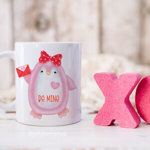 Op de afbeelding: Een witte keramische mok met een roze pingu&iuml;n die een rode strik draagt en een rode envelop met een hart vasthoudt. De pingu&iuml;n heeft de woorden "be mine" op zijn borst. De mok staat op een wit houten oppervlak met roze schuimletters die "XO" ervoor spellen.