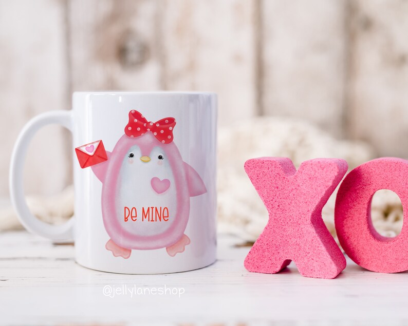 Op de afbeelding: Een witte keramische mok met een roze pingu&iuml;n die een rode strik draagt en een rode envelop met een hart vasthoudt. De pingu&iuml;n heeft de woorden "be mine" op zijn borst. De mok staat op een wit houten oppervlak met roze schuimletters die "XO" ervoor spellen.