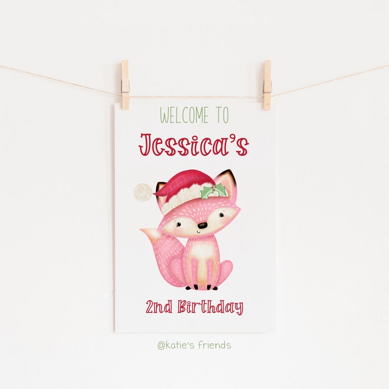 May include: A white card with a pink fox wearing a Santa hat, with the text "WELCOME TO Jessica's 2nd BIRTHDAY". The card is hanging from a string with wooden clothespins.