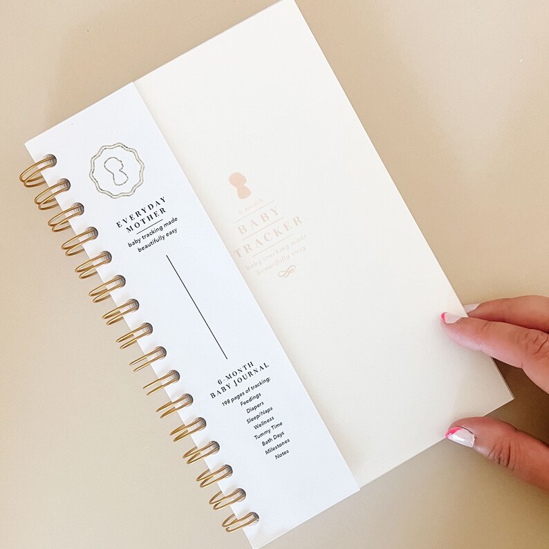 Image of a daily baby tracking notebook with grey cover and gold spiral binding. The cover has a gold logo. The 5.5x8.5 inch notebook,  is made in New York. Designed by a NICU mom, it facilitates tracking of feeds, diapers, sleep, and notes for baby.