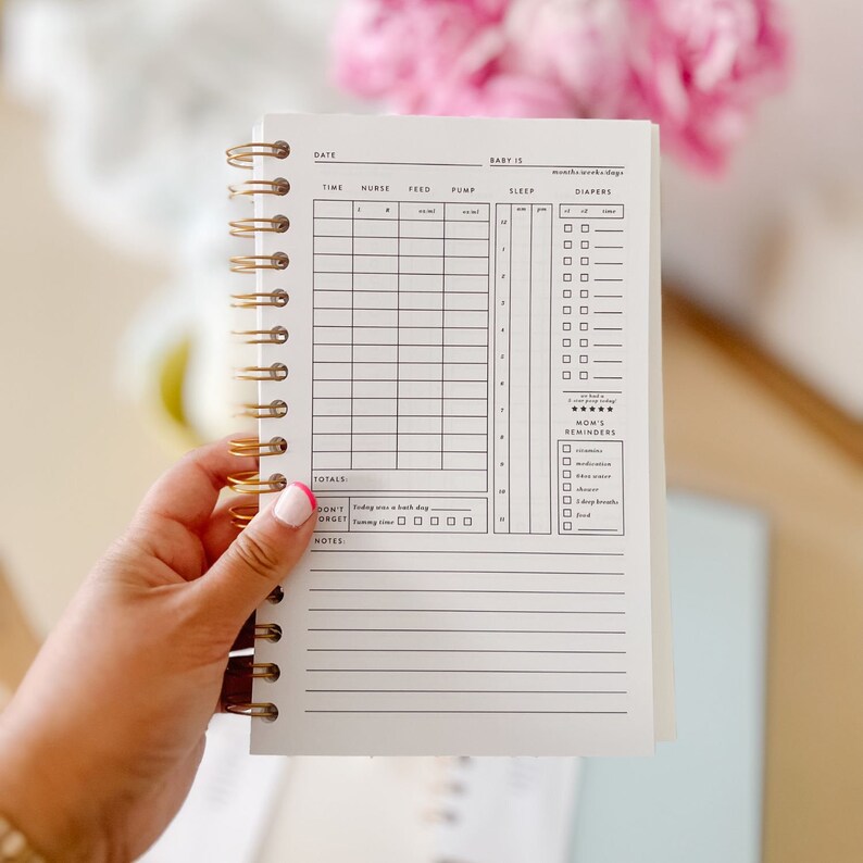 An open daily baby tracking notebook displaying a user-friendly template page for monitoring baby activities. The page features organized sections for recording feeds, diaper changes, sleep schedules, milestones, and additional notes.