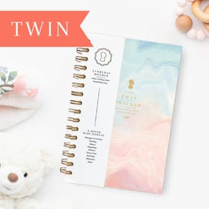 May include: A spiral-bound baby journal with the word "TWIN" in a red banner. The journal has a white cover with gold spiral binding and the text "EVERYDAY MOTHER" and "TWIN TRACKER". The journal's cover has a watercolor design in shades of blue and pink.