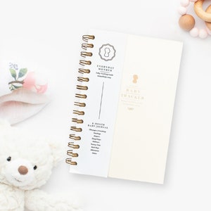 Image of a daily baby tracking notebook with grey cover and gold spiral binding. The cover has a gold logo. The 5.5x8.5 inch notebook,  is made in New York. Designed by a NICU mom, it facilitates tracking of feeds, diapers, sleep, and notes for baby.