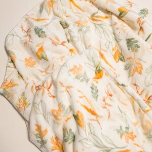 May include: A white fleece blanket with a pattern of orange and green leaves. The blanket is soft and cozy, perfect for snuggling up on the couch.