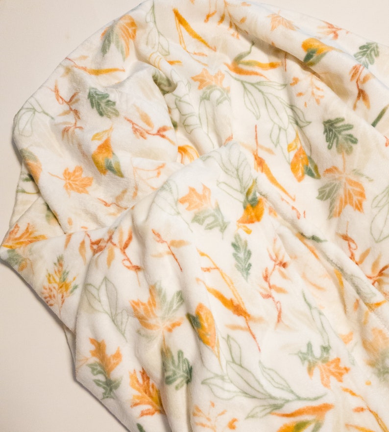 May include: A white fleece blanket with a pattern of orange and green leaves. The blanket is soft and cozy, perfect for snuggling up on the couch.