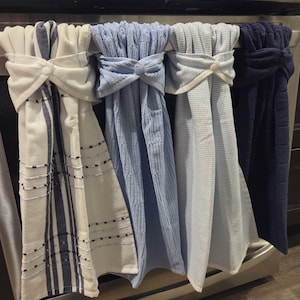 May include: A set of four kitchen towels in various shades of blue and white, hanging on a stainless steel oven. The towels are adorned with decorative knots and bows. The first towel has vertical navy stripes, while the others are solid or striped.
