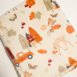 May include: A cream-colored fleece blanket with a fall-themed print. The print features orange, red, and brown deer, pumpkins, trees, leaves, and a red truck.