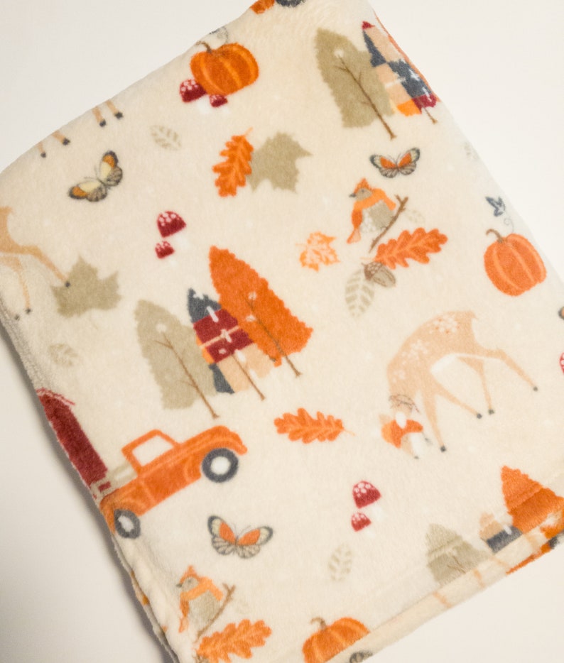 May include: A cream-colored fleece blanket with a fall-themed print. The print features orange, red, and brown deer, pumpkins, trees, leaves, and a red truck.
