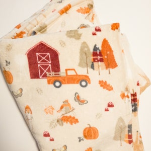May include: A cream-colored fleece blanket with a fall-themed print. The print features a red barn, a red truck, pumpkins, leaves, and birds.